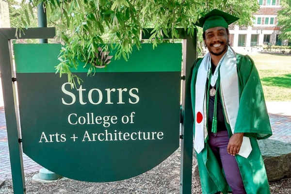 photo of recent graduate in cap and gown in front of the storrs sign