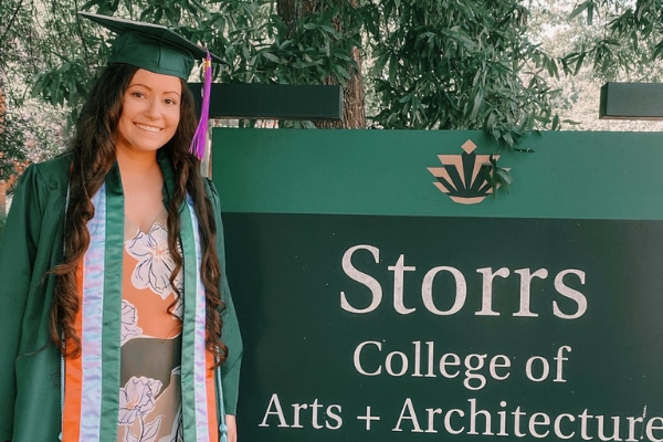 photo of recent graduate in cap and gown in front of the storrs sign