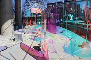 colorful light installation of transparent curved forms on sidewalk next to light rail line