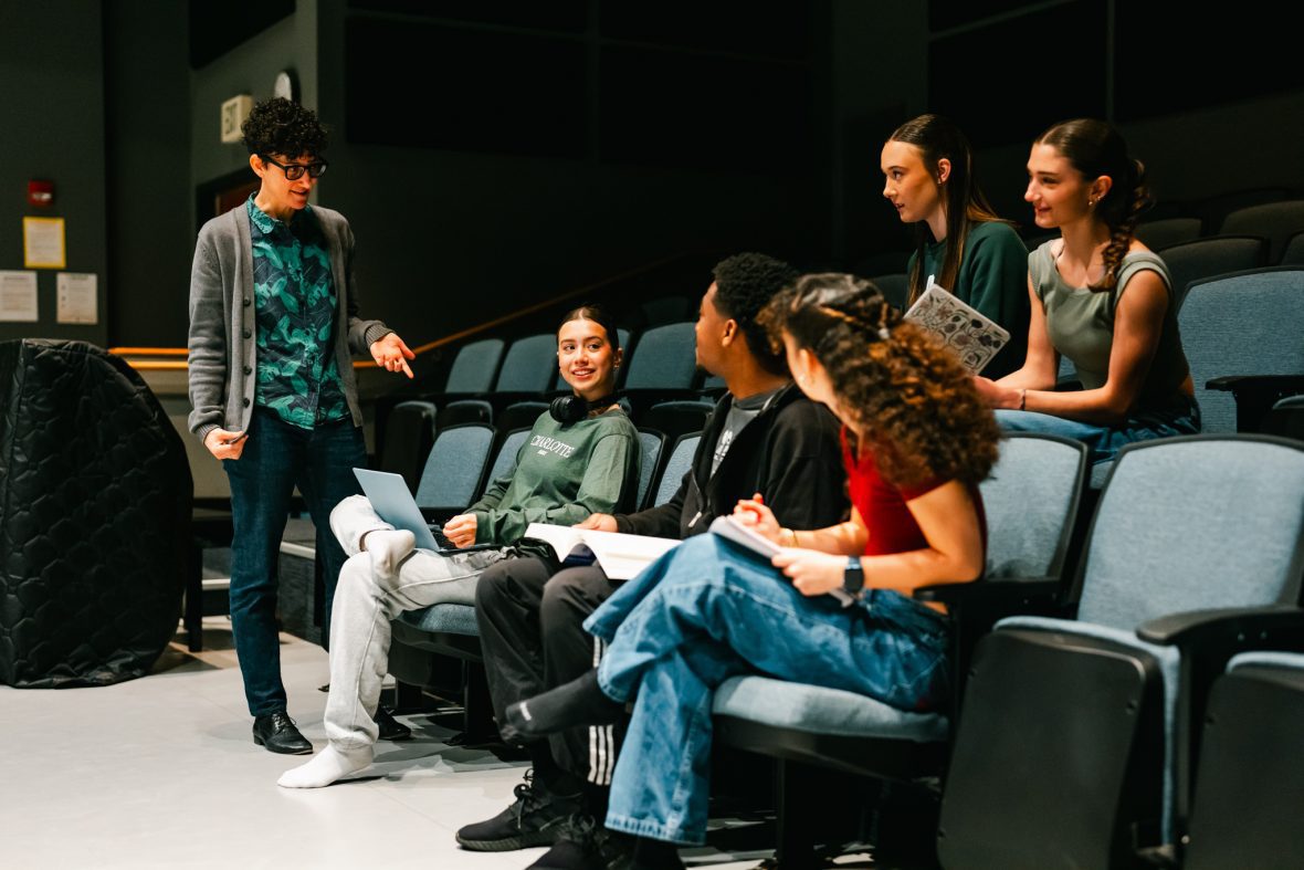 Dance students sitting in the chairs in the studio, listening to professor with notebooks in their hands