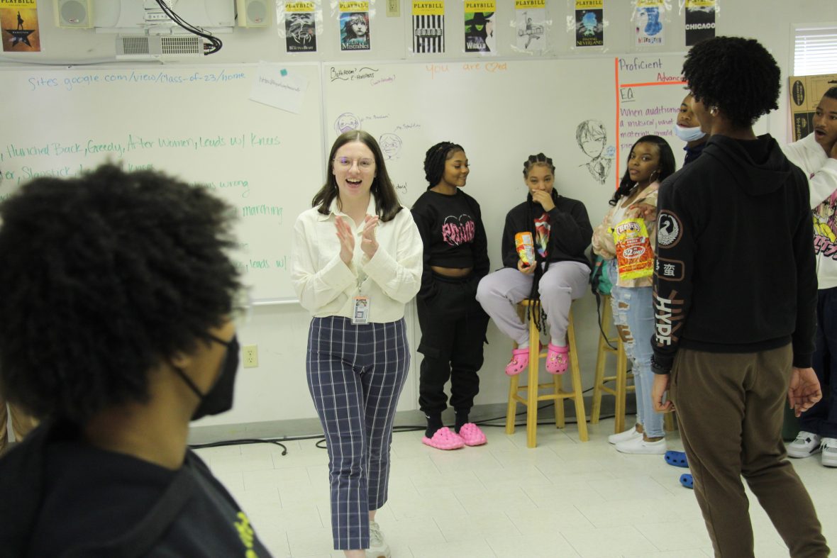 Young woman in a theatre classroom in a public school teaching a theatre class. She is smiling with her hands clapped together.