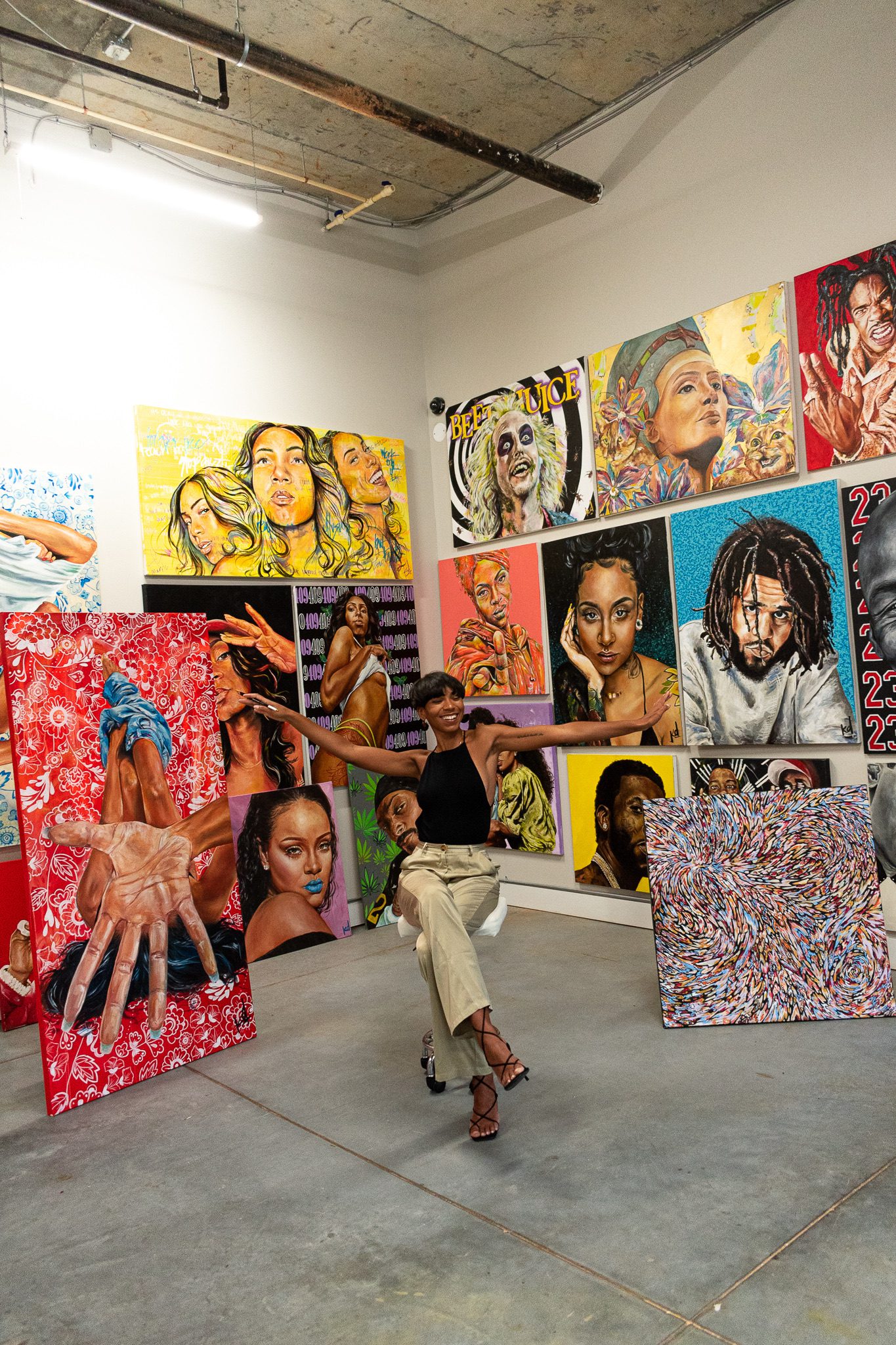 art alumna sitting in an industrial space surrounded by her paintings with arms outstretched