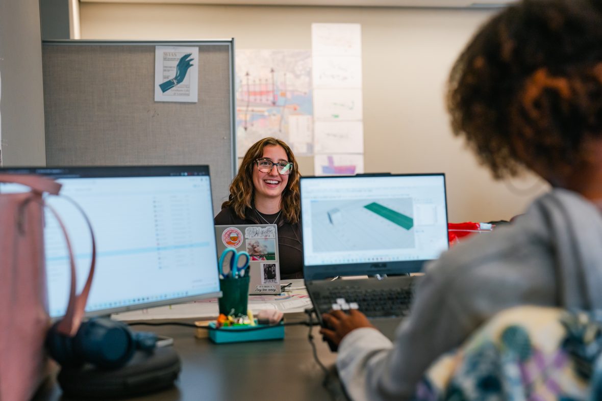 student smiling in the background with two laptop screens in front