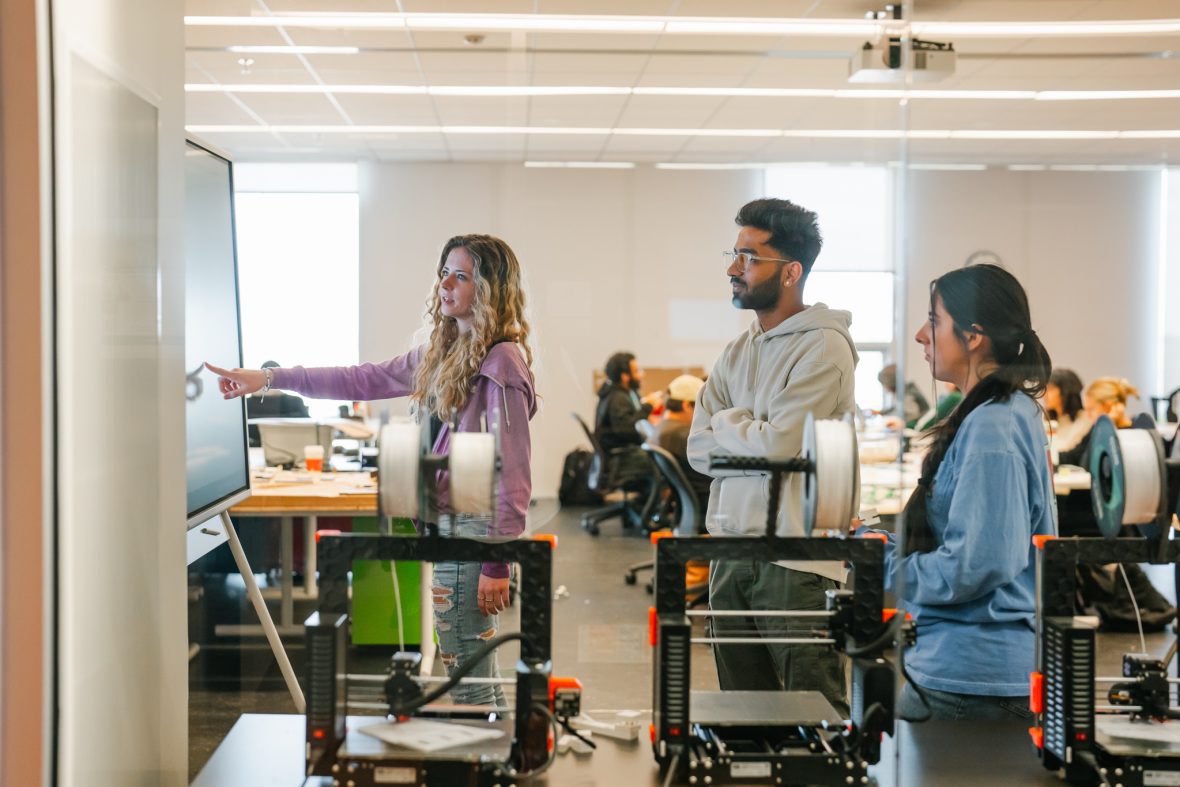 three architecture students working on a project together looking at a monitor with machinery in front of them