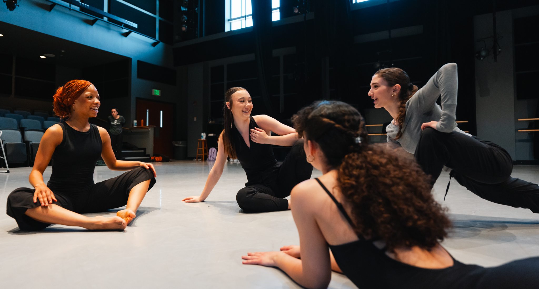 Four dance students sitting on the ground of the studio, stretching, talking and smiling