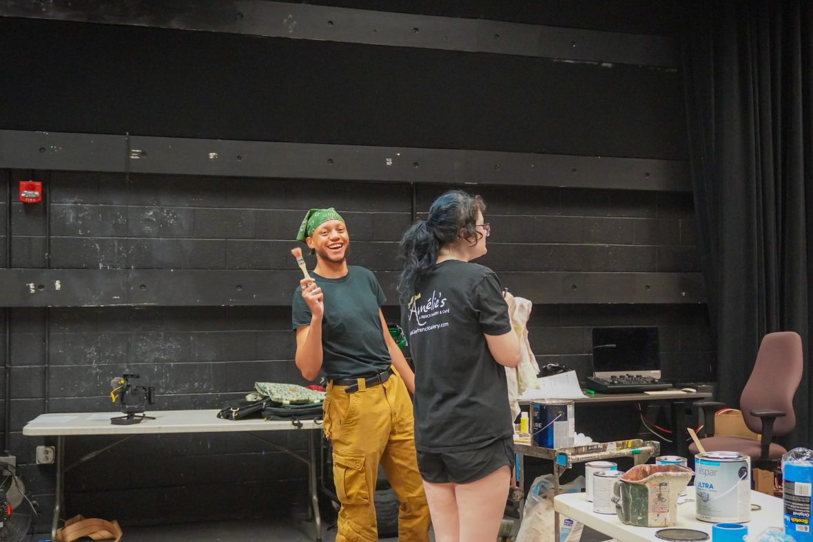 Two students in the black box theater working on a set design, one holding a paint brush