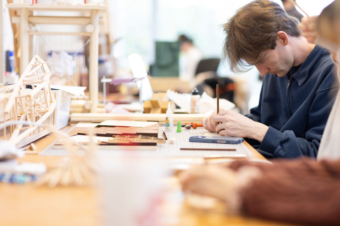 male architecture student writing in a notebook with models behind him