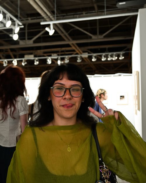 woman in a green blouse and glasses giving the peace sign in an art gallery