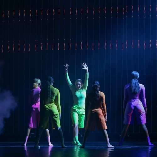 group of dancers in colorful costumes in a darkly lit stage with red LED lights and moke