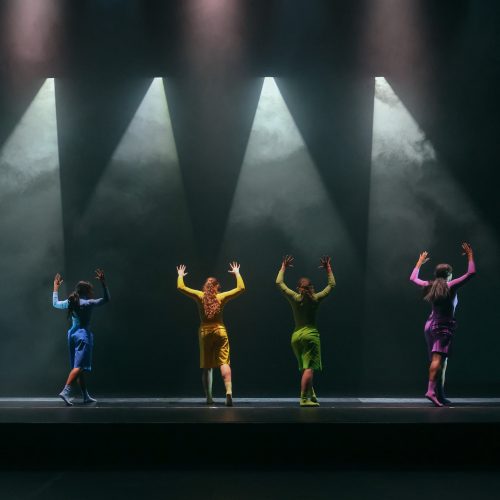 Four dancers in colorful costumes in a darkly lit stage with spotlights and smoke