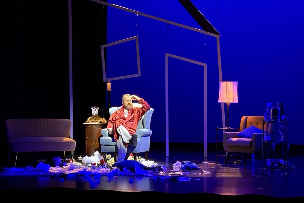 A scene from the play "Wink," with an actor sitting in an arm chair, wearing his bathrobe.