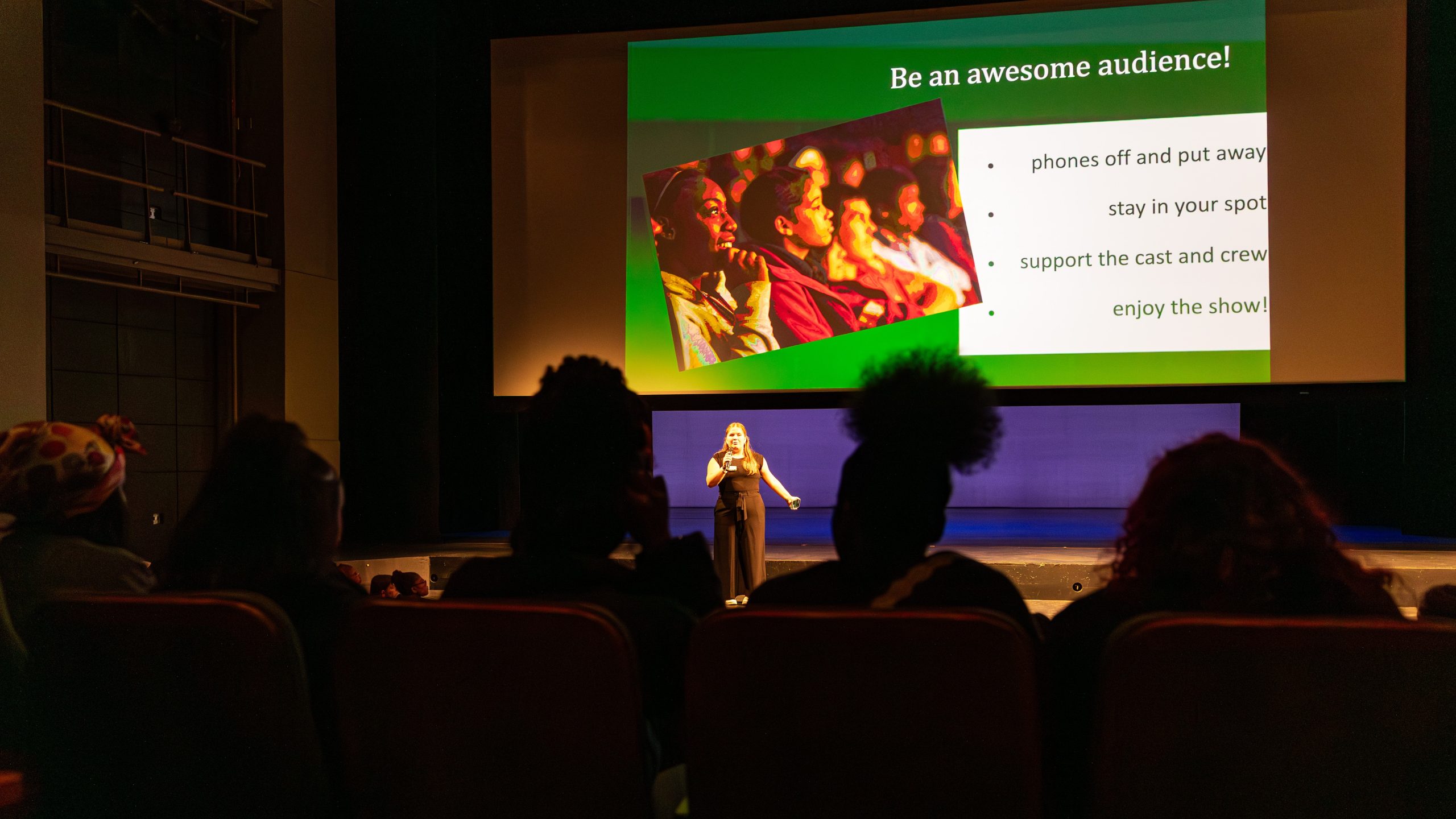 UNC Charlotte student Maddie Worsley introduces the concert, standing on stage, with projection behind her reading Be and awesome audience!